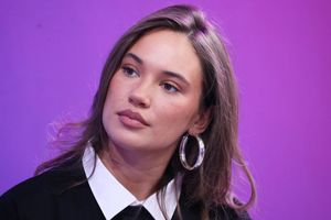 Młoda kobieta z długimi włosami, w czarnej koszuli i dużych kolczykach, na purpurowym tle.