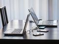 Na stole stoją dwa laptopy i kilka smartfonów, wszystko na tle jasnych zasłon.