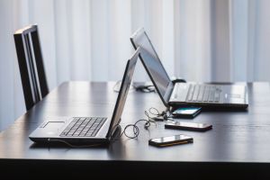 Na stole stoją dwa laptopy i kilka smartfonów, wszystko na tle jasnych zasłon.