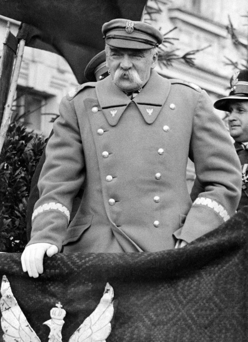 Mężczyzna w wojskowym mundurze, z wąsami, trzyma flagę z orłem. Na tle zieleni i innych osób.