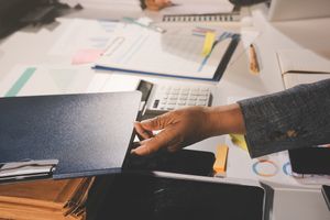 Na biurku widać rękę sięgającą po teczkę, wokół dokumenty i urządzenia biurowe.