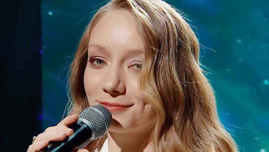 Młoda kobieta z mikrofonem, uśmiechająca się, na tle niebieskiego oświetlenia.