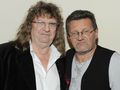 Dwaj mężczyźni w casualowych ubraniach, stoją blisko siebie z poważnymi minami na tle jasnej ściany.