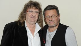 Dwaj mężczyźni w casualowych ubraniach, stoją blisko siebie z poważnymi minami na tle jasnej ściany.