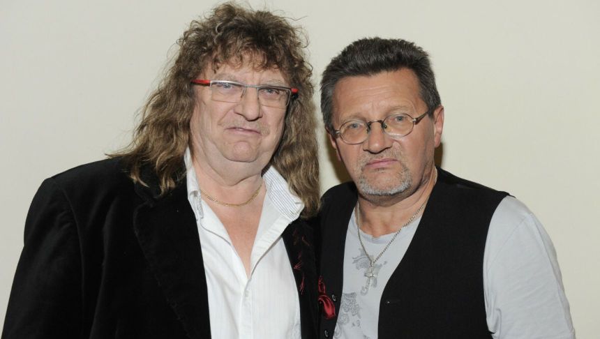 Dwaj mężczyźni w casualowych ubraniach, stoją blisko siebie z poważnymi minami na tle jasnej ściany.