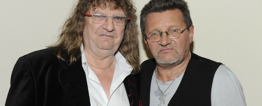 Dwaj mężczyźni w casualowych ubraniach, stoją blisko siebie z poważnymi minami na tle jasnej ściany.