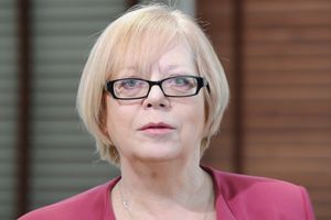 Kobieta z blond włosami w różowej marynarce, stoi na tle drewnianej ściany.