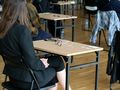 Osoba w czarnym garniturze siedzi przy biurku, wokół innych uczestników w tle.