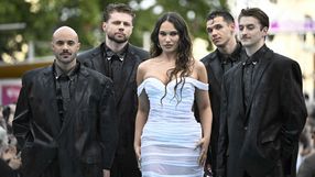Czterej mężczyźni w czarnych garniturach i jedna kobieta w białej sukni na tle publiki.