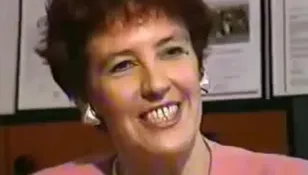 Uśmiechnięta kobieta w różowej bluzce, z krótkimi włosami, na tle dokumentów.