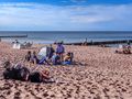 Ludzie odpoczywają na plaży, słońce świeci, woda i piasek w tle. Atmosfera relaksu i zabawy.