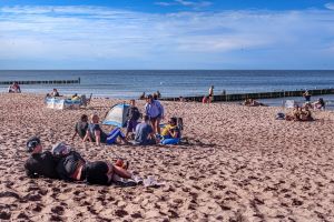 Ludzie odpoczywają na plaży, słońce świeci, woda i piasek w tle. Atmosfera relaksu i zabawy.