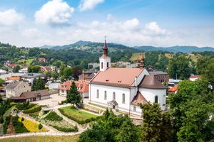Widok na biały kościół z czerwonym dachem w otoczeniu zielonych wzgórz i domów.