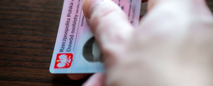 Na zdjęciu widoczna ręka trzymająca polski dowód osobisty na drewnianym stole.