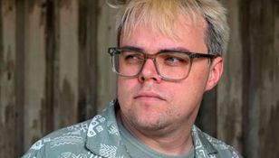 Mężczyzna z okularami, z blond włosami, patrzący w bok na tle drewnianej ściany.