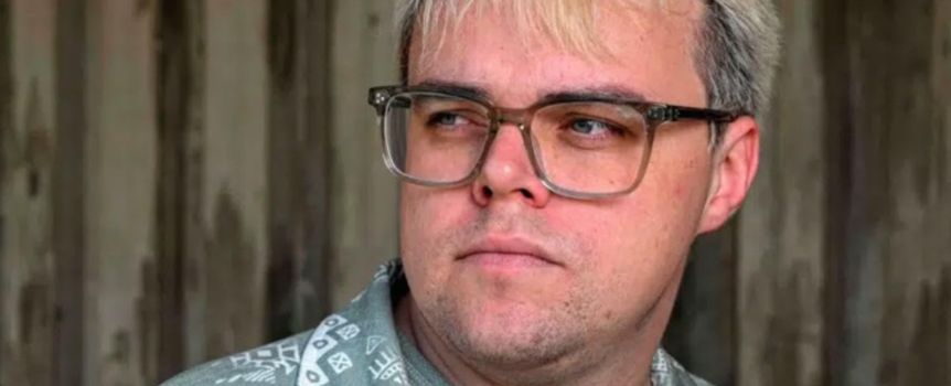 Mężczyzna z okularami, z blond włosami, patrzący w bok na tle drewnianej ściany.