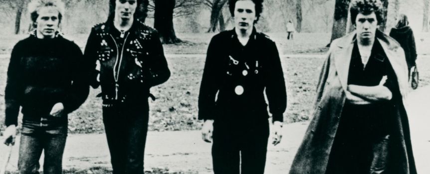 Czterech mężczyzn w stylu punk, idących w parku, w czarno-białym zdjęciu z lat 70. XX wieku.