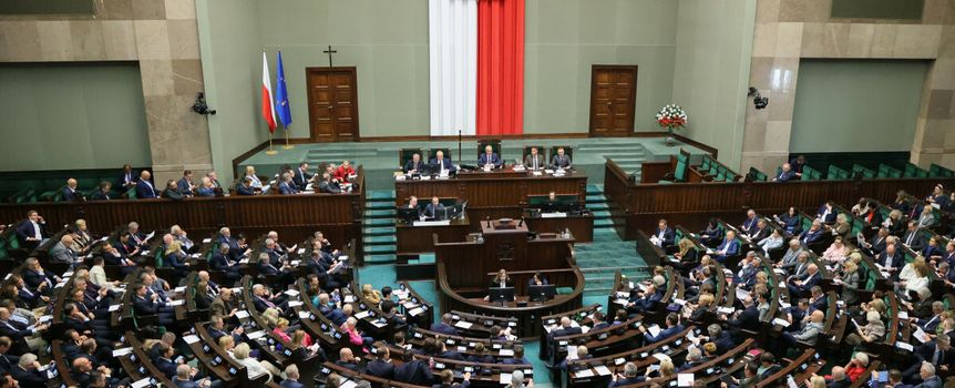 Zgromadzenie w Sejmie, z flagą Polski i orłem na ścianie, wielu przedstawicieli w sali.