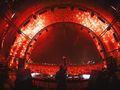 Tłum festiwalu muzycznego, DJ na scenie, jasne światła, efekty pirotechniczne w tle.