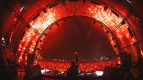 Tłum festiwalu muzycznego, DJ na scenie, jasne światła, efekty pirotechniczne w tle.