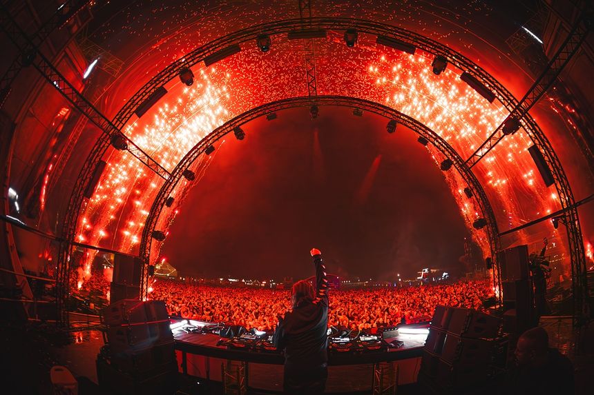 Tłum festiwalu muzycznego, DJ na scenie, jasne światła, efekty pirotechniczne w tle.