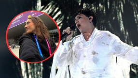 Kobieta w czarnej bluzie z identyfikatorem, obok artysta w białej kreacji śpiewający na scenie.