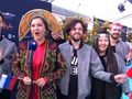 Grupa ludzi z flagami, ubrana w srebrne kurtki, w kolorowych strojach folklorystycznych.