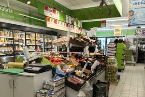 Wnętrze sklepu: półki z warzywami, stojaki z napojami, kasa i wywieszki z cenami.