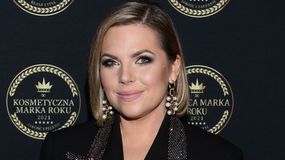 Aleksandra Kwaśniewska z krótkimi włosami w eleganckim stroju, uśmiechająca się na tle ciemnej plakaty.