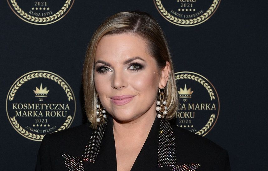 Aleksandra Kwaśniewska z krótkimi włosami w eleganckim stroju, uśmiechająca się na tle ciemnej plakaty.
