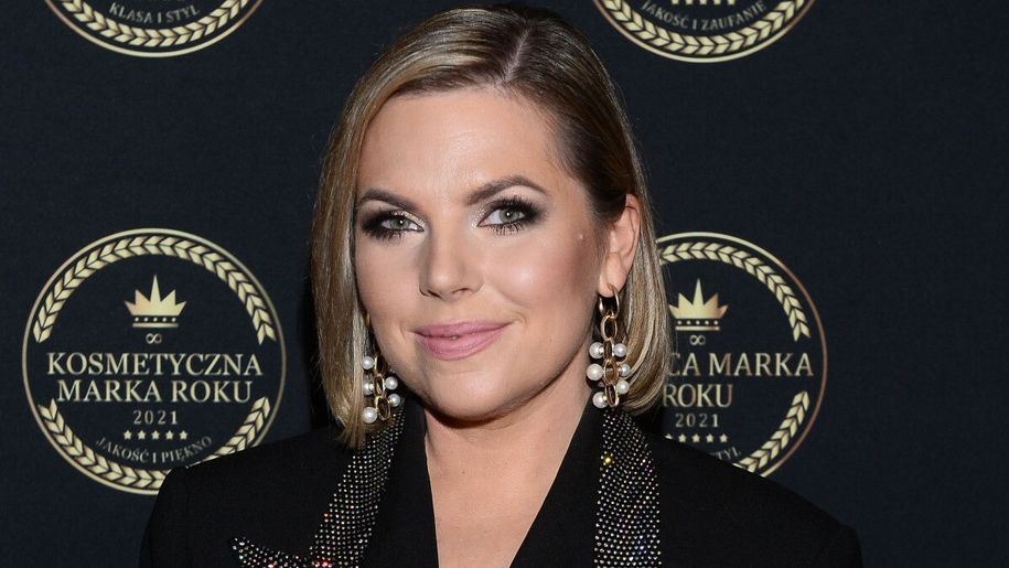 Aleksandra Kwaśniewska z krótkimi włosami w eleganckim stroju, uśmiechająca się na tle ciemnej plakaty.