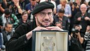 Mężczyzna w beretach uśmiecha się, trzymając nagrodę. W tle grupka fotografów.