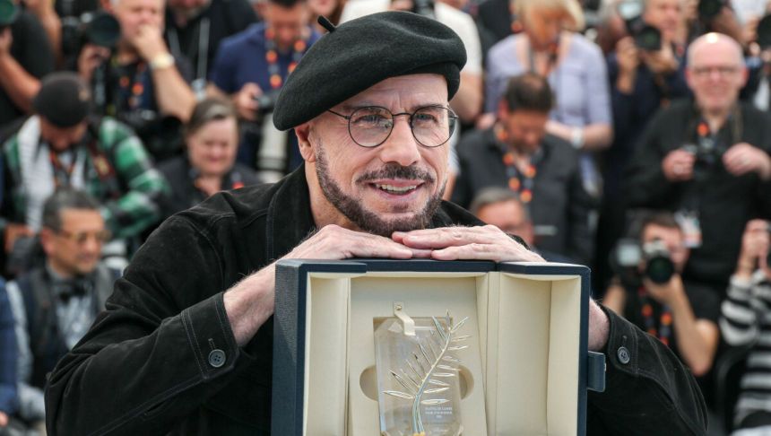 Mężczyzna w beretach uśmiecha się, trzymając nagrodę. W tle grupka fotografów.