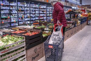 Kobieta w czerwonej kurtce przy stoisku z warzywami w supermarkecie, obok wózek na zakupy.