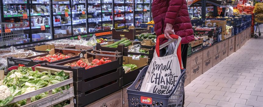 Kobieta w czerwonej kurtce przy stoisku z warzywami w supermarkecie, obok wózek na zakupy.