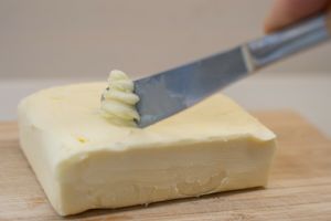 Na drewnianej desce leży kostka masła, na którą nakładany jest kawałek masła nożem.