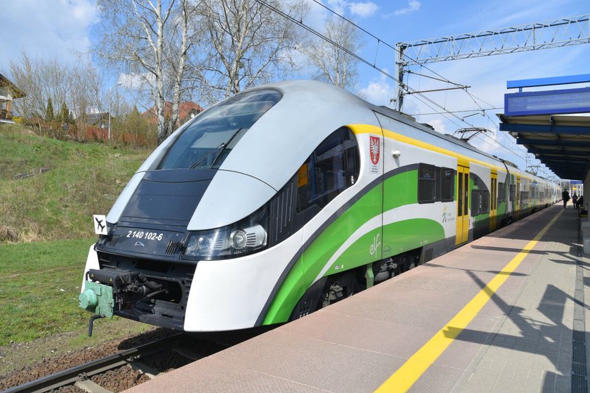 Pociąg elektryczny stoi na peronie, w tle niebo z chmurami i drzewa.