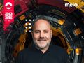 DJ Kris z uśmiechem w ciemnej koszulce na tle technologicznego, kolorowego wzoru.
