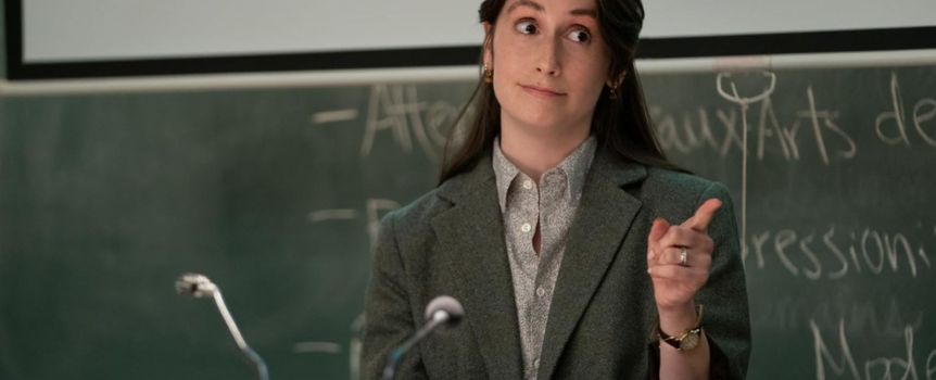 Kobieta w marynarce, wskazująca palcem, stoi przed tablicą w sali wykładowej.