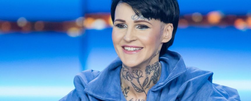 Na zdjęciu uśmiechnięta kobieta w niebieskiej bluzie, z tatuażami na szyi i dekolcie.