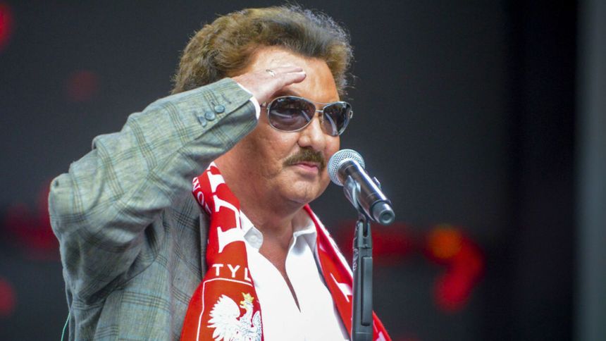 Mężczyzna w okularach przeciwsłonecznych, w rozpinanej koszuli, salutuje przy mikrofonie.