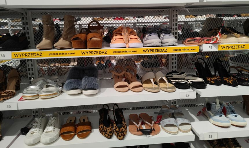 Na zdjęciu półki z różnorodnymi obuwiem na wyprzedaży, w tym sandały i sneakersy.