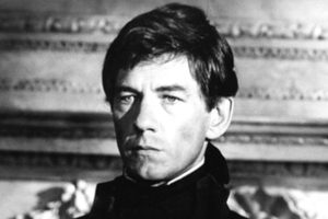 Ian McKellen w ciemnym stroju, poważny wyraz twarzy, klasyczne tło. Portret w czerni i bieli.