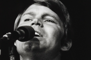 Glen Campbell śpiewa do mikrofonu, w tle ciemna scena. Czarno-biała fotografia.
