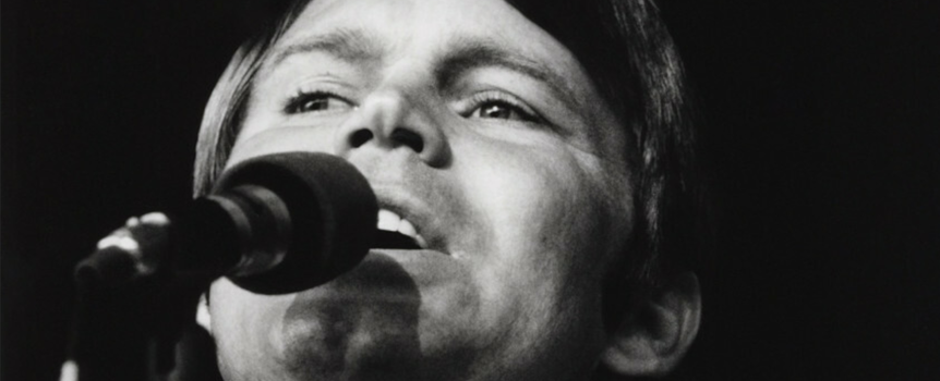 Glen Campbell śpiewa do mikrofonu, w tle ciemna scena. Czarno-biała fotografia.