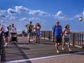 Spacerowicze na drewnianym molo, plaża w tle, błękitne niebo z chmurami. Brak rozpoznawalnych polityków.