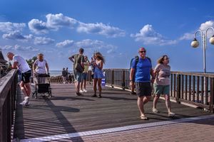 Spacerowicze na drewnianym molo, plaża w tle, błękitne niebo z chmurami. Brak rozpoznawalnych polityków.