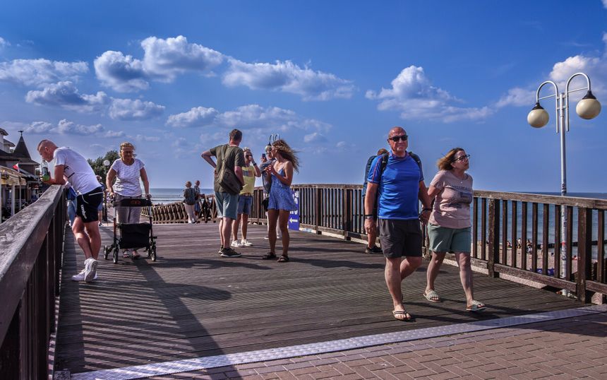Spacerowicze na drewnianym molo, plaża w tle, błękitne niebo z chmurami. Brak rozpoznawalnych polityków.