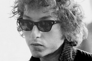 Bob Dylan z kręconymi włosami w okularach przeciwsłonecznych, na tle jaśniejszej ściany.