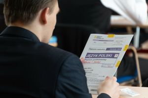 Uczeń w garniturze trzyma arkusz egzaminu maturalnego z języka polskiego.